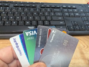 A hand holding several credit and debit cards, including Capital One, Visa, and HealthEquity, in front of a black computer keyboard on a wooden desk.